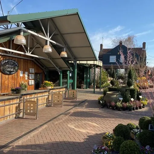 Outdoor garden centre courtyard with wooden buildings, green canopy roof, hanging lights, flowering plants, and a brick farmhouse visible behind.