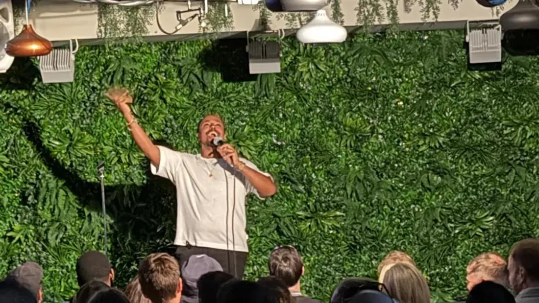 Performer holding microphone on stage against green foliage backdrop, addressing audience seated below.