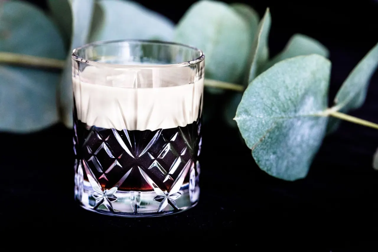 Cut glass tumbler with dark spirit and cream layer beside eucalyptus leaves against black background.