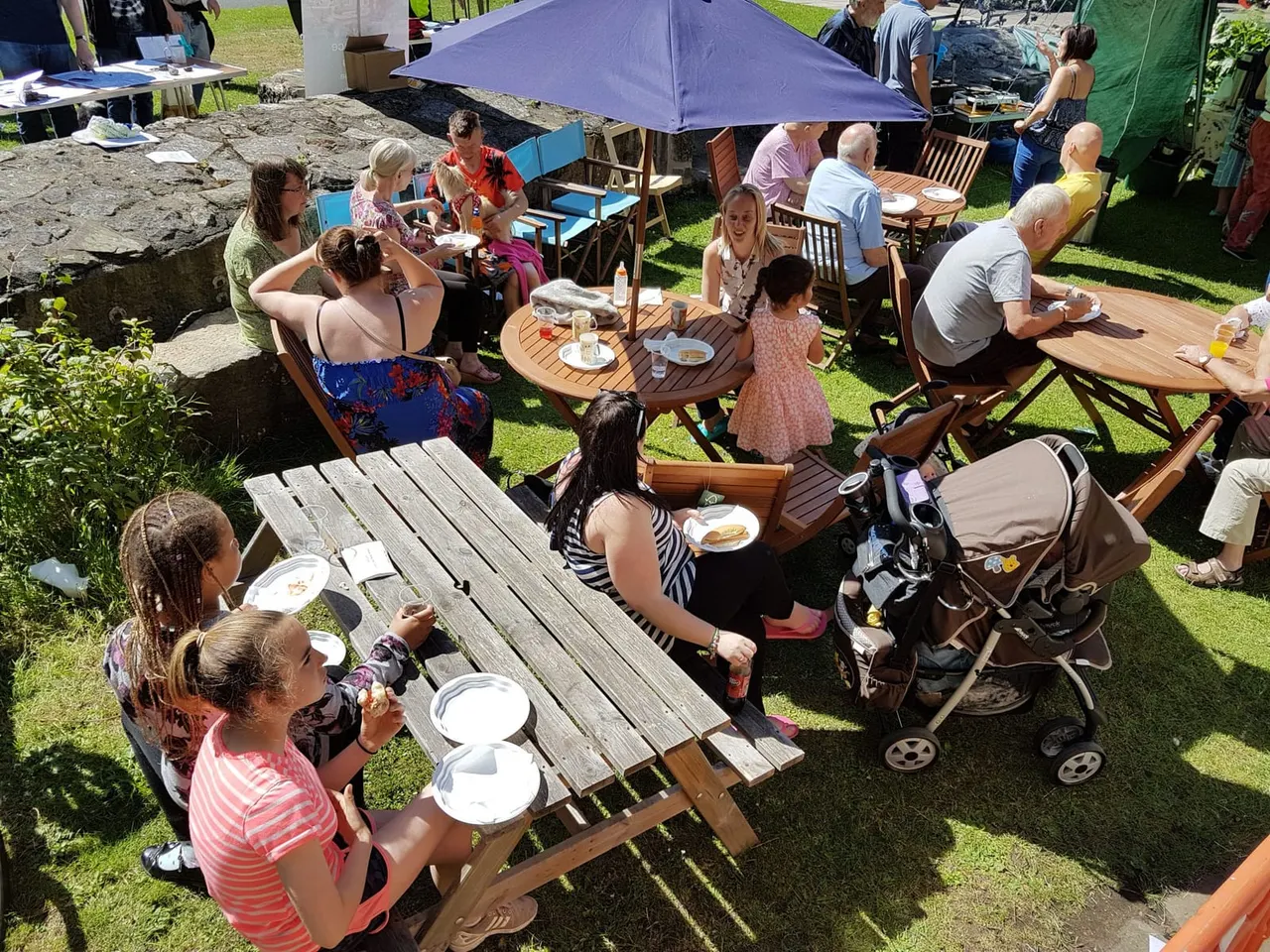 Families and visitors of mixed ages enjoying outdoor picnic tables and meals on a sunny grass field, with umbrellas and camping chairs scattered throughout.