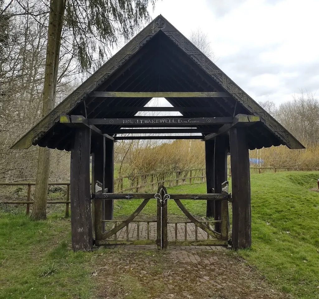 A traditional wooden pavilion shelter with ornate decorative gates in a rural countryside setting with bare trees and open grassland.
