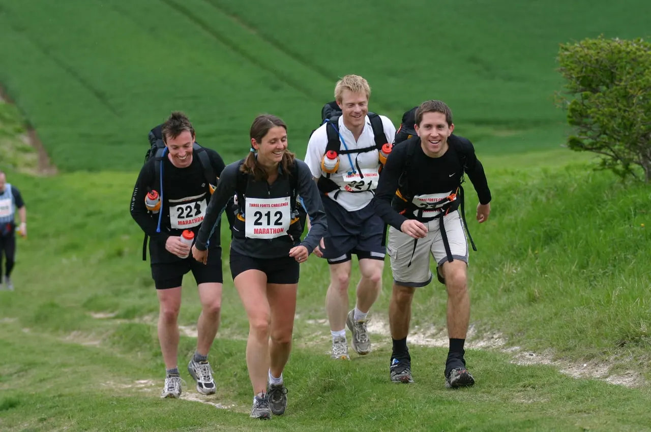 Four runners with race bibs jogging together along a grassy hillside path during a marathon event.