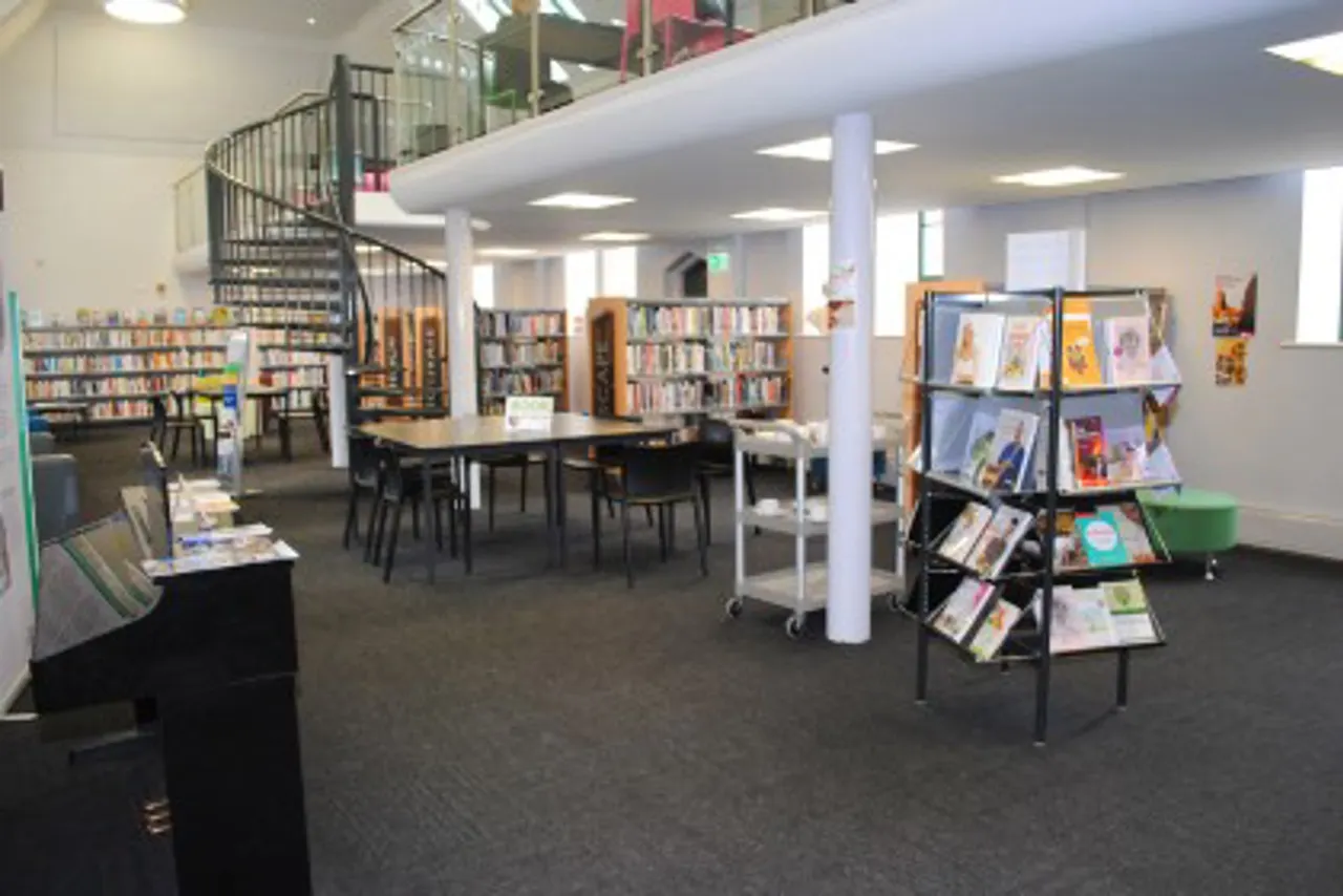 Library interior with bookshelves, display stands, reading tables, spiral staircase and seating areas.