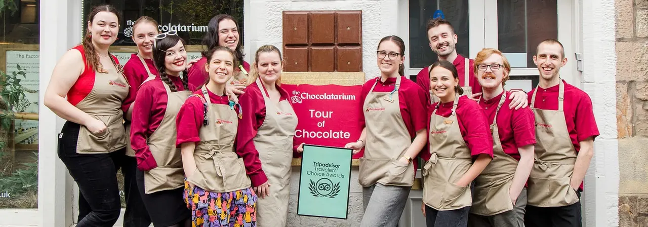 A team of staff members in red and beige uniforms pose together outside the Chocolatarium, holding a "Tour of Chocolate" banner and a TripAdvisor Travellers' Choice Awards sign.