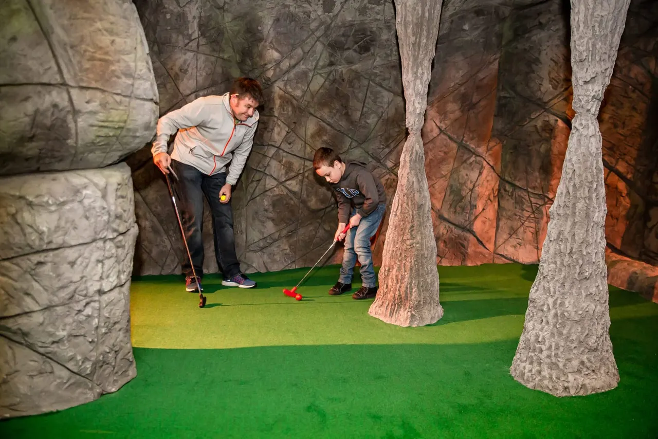An adult and child playing indoor adventure golf with oversized tree trunk obstacles and a forest backdrop.