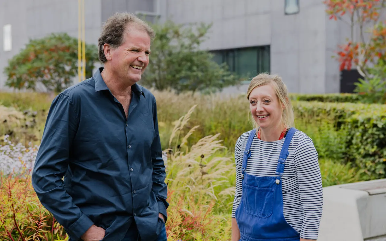 Two people smiling in a contemporary garden with ornamental grasses and planted beds, modern building behind.