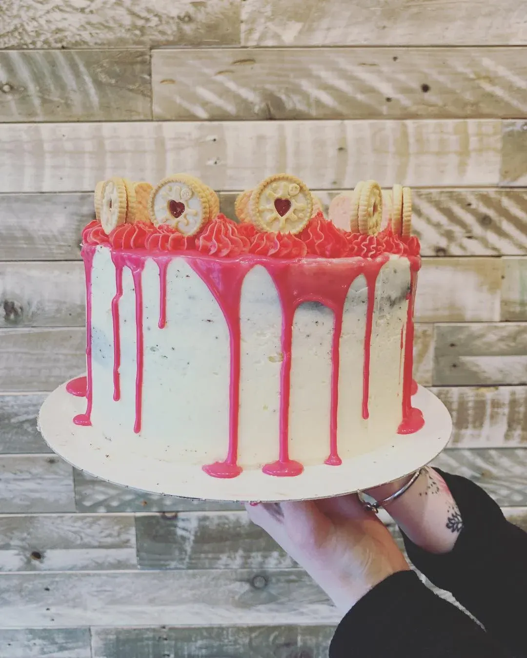Person holding a pink and white drip cake decorated with biscuits and heart jam centres against a wooden backdrop.