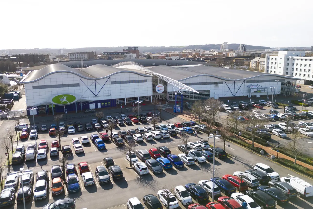 Modern leisure centre with distinctive wave-shaped roof and glazed facade, surrounded by extensive car park with numerous vehicles and urban skyline beyond.