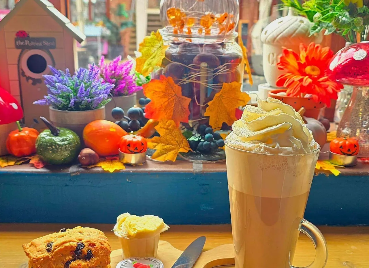 A glass of iced coffee with cream topping sits on a table with a scone, in front of an autumn-themed display featuring flowers, pumpkins, and decorative leaves.