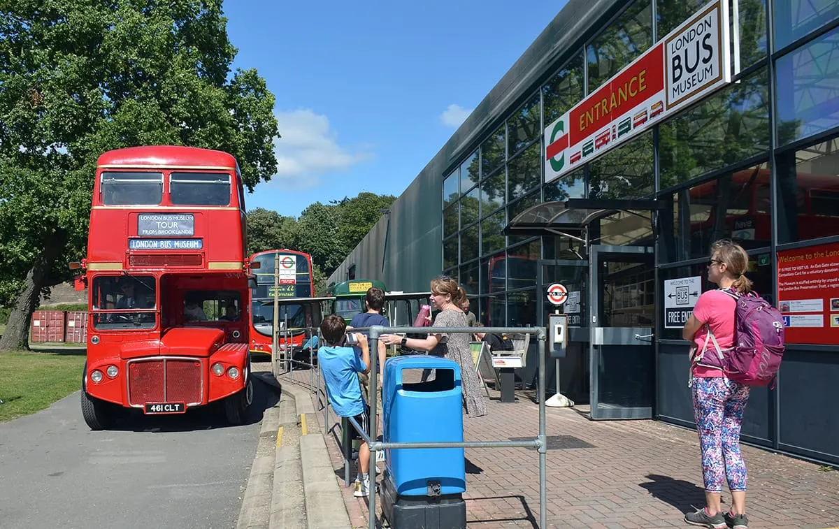 A red London Routemaster bus parked outside the museum entrance with visitors and children queuing nearby on a sunny day.