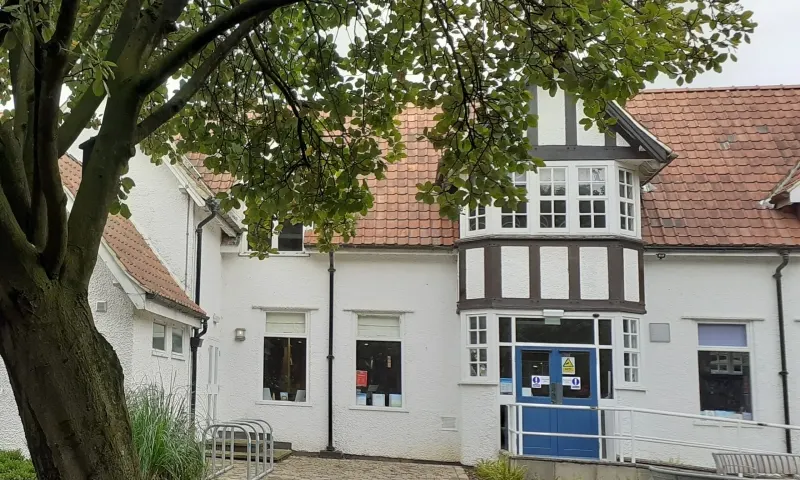 Historic white-rendered building with half-timbered upper storey and terracotta roof, large mature tree in foreground, blue entrance door visible.