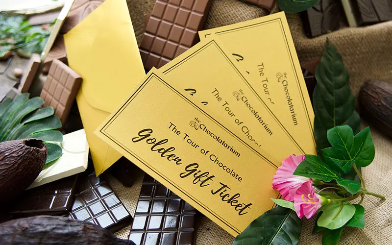 Golden gift vouchers and chocolate bars arranged with green foliage and a pink flower on neutral fabric.
