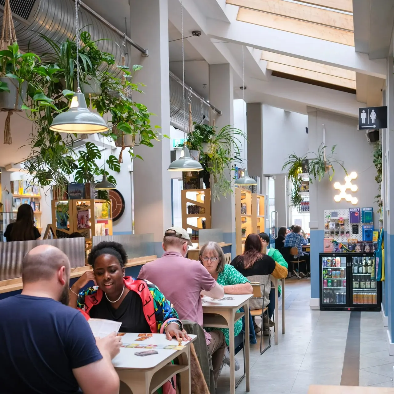 A bright, modern café interior with visitors seated at tables playing board games beneath hanging plants and pendant lights.