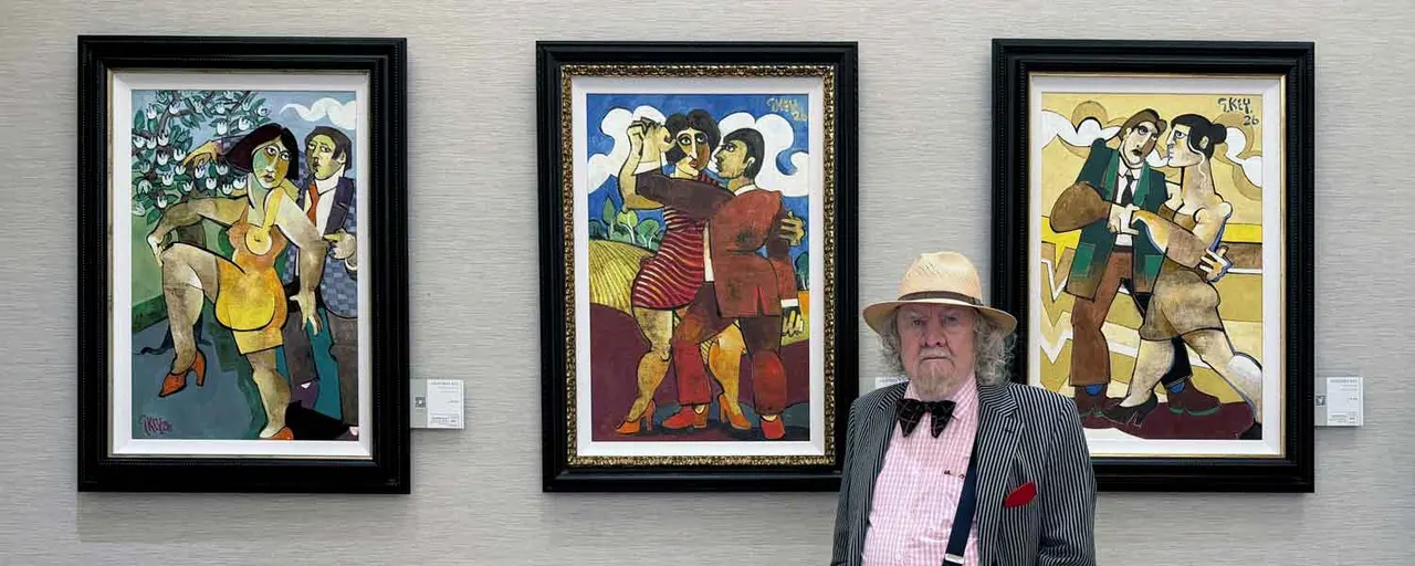 An elderly man in a straw hat and formal attire stands before three colourful framed figurative artworks depicting people in various scenes.