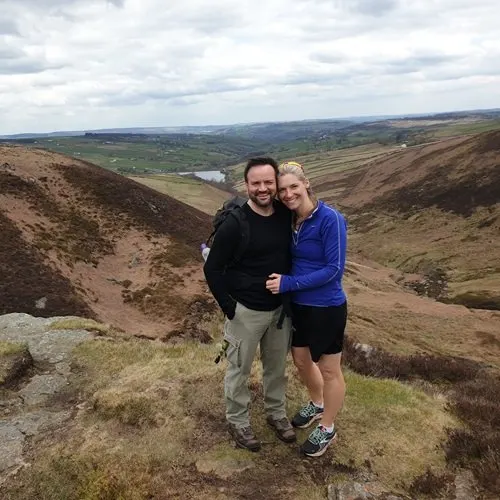 Two people posing on moorland with rolling hills and valley views beyond.