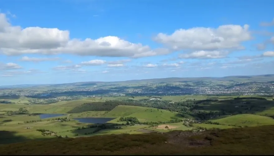 Aerial view of rolling green countryside with fields, woodland, and lakes stretching to distant hills under blue sky.