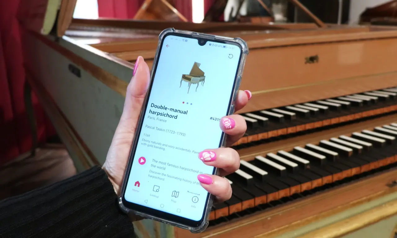 Hand holding a smartphone displaying information about a harpsichord, with a grand piano visible in the background.