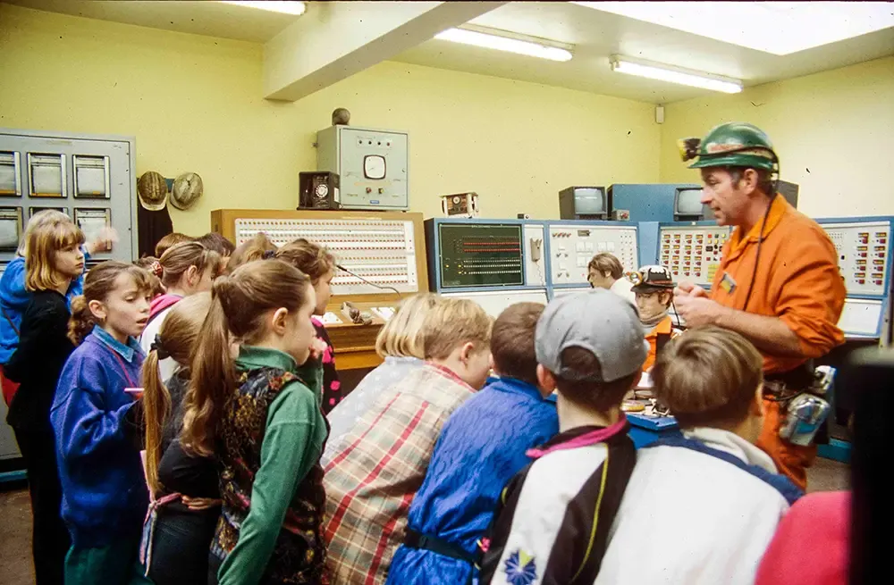 Children gather around an adult guide in a vintage electronics laboratory filled with vintage computer equipment and control panels.