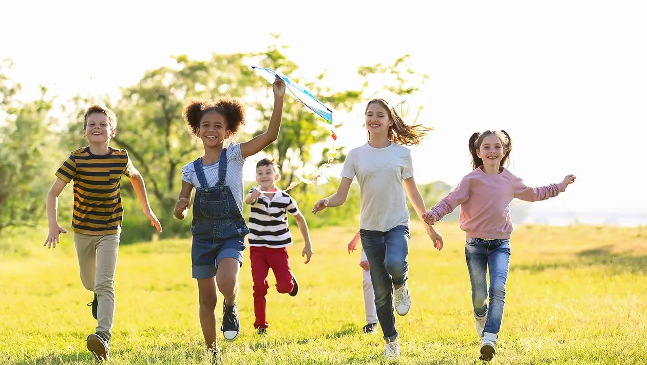 A group of five children running through a sunny grassy field, one holding a blue kite above her head.