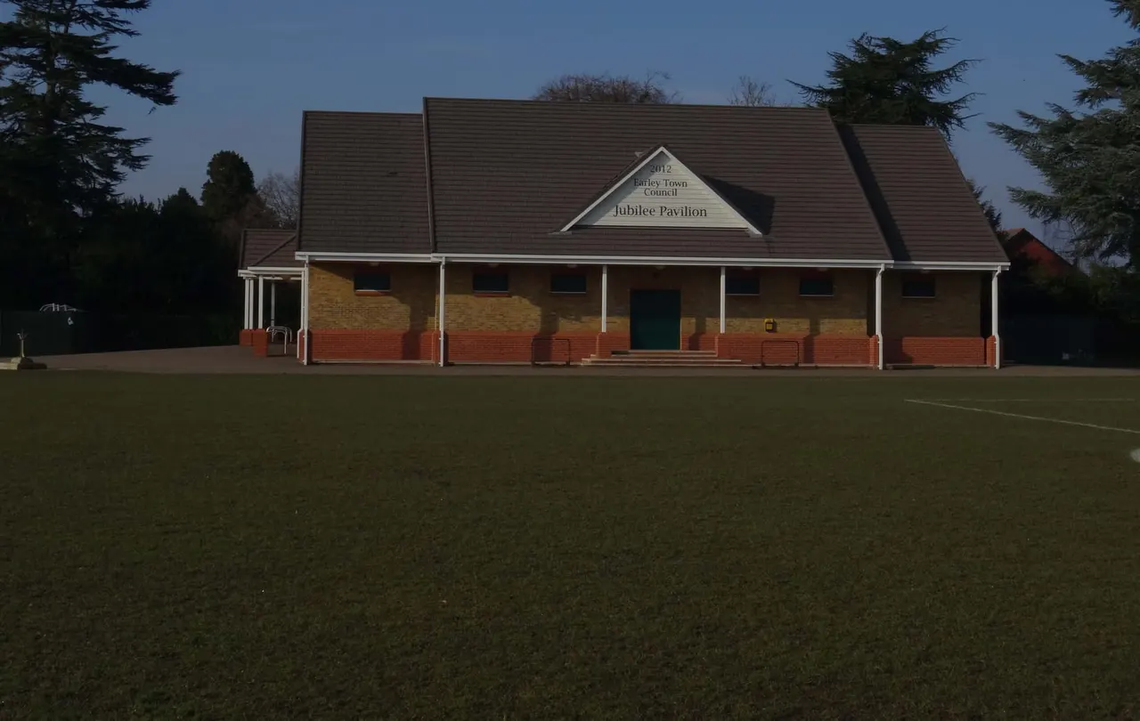 Brick pavilion building with covered porch set on open grass field, surrounded by conifer trees.