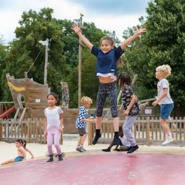 Children playing on a trampoline at an outdoor play area with a wooden pirate ship structure in the background.