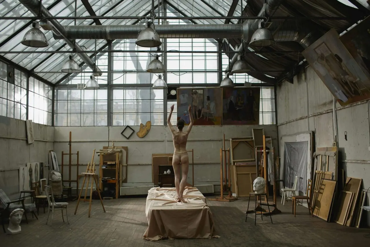Artist posing as a live model in an industrial studio space with easels, skylight, and classical busts.