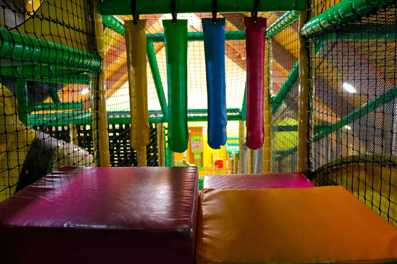 Interior of a children's play structure with coloured foam pads, netting, and hanging cylindrical obstacles.