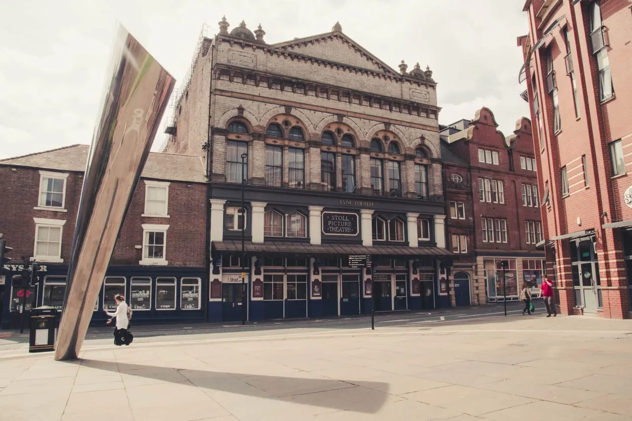 Historic Victorian theatre building with ornate stonework and arched windows on a town square, with pedestrians and modern sculpture nearby.