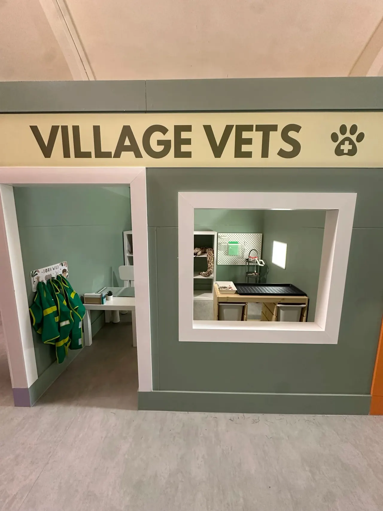A child-sized play veterinary clinic with green walls, featuring a service window and open door entrance with green uniforms hanging inside.