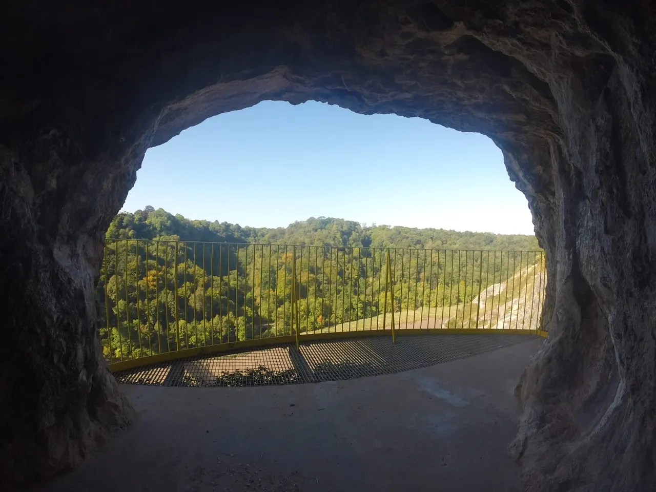 View from inside a cave or tunnel looking out towards a metal-railed viewing platform overlooking forested countryside.