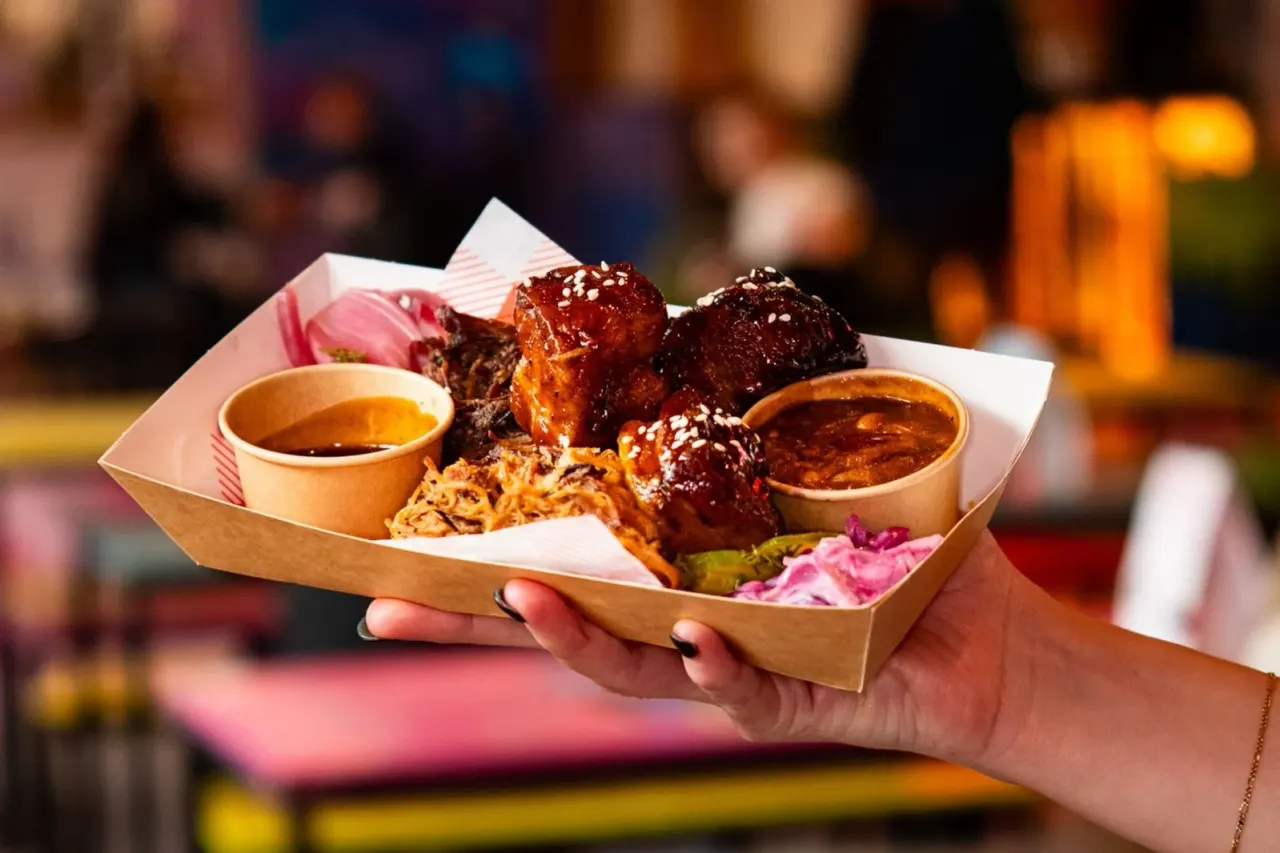 Hand holding cardboard tray of glazed meat balls with dipping sauces and noodles at outdoor food event.