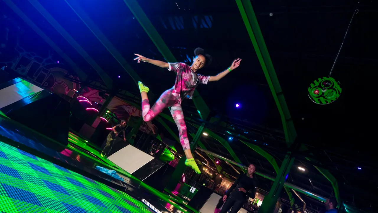 Person in neon pink outfit jumping on a trampoline in a brightly lit indoor arena with green and blue lighting.