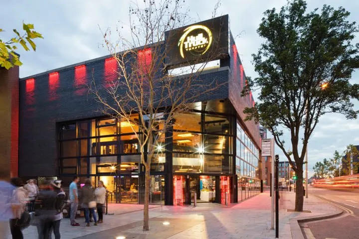 Modern theatre building with illuminated signage, evening street scene with pedestrians and trees outside.