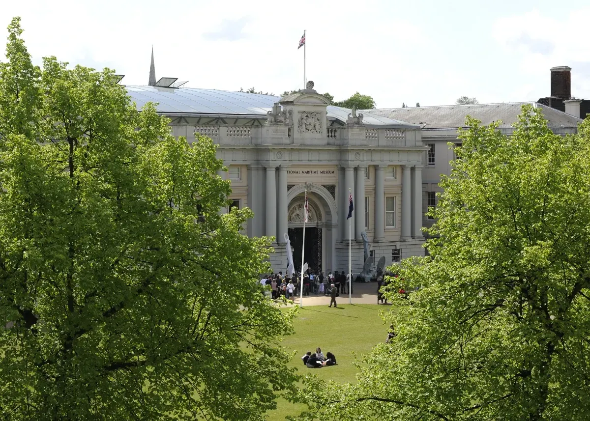 Grand neoclassical museum building with columns and flags, framed by green trees, with visitors on the lawn below.