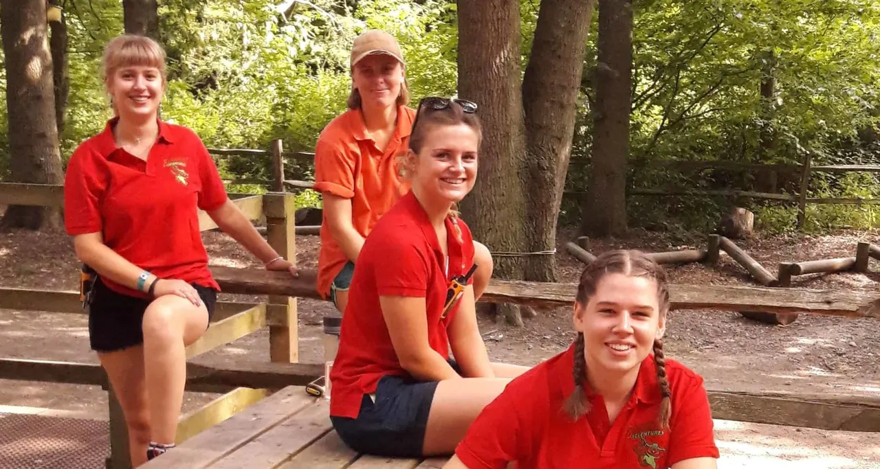 Four staff members in red uniforms sitting on wooden platforms in a woodland adventure activity area.