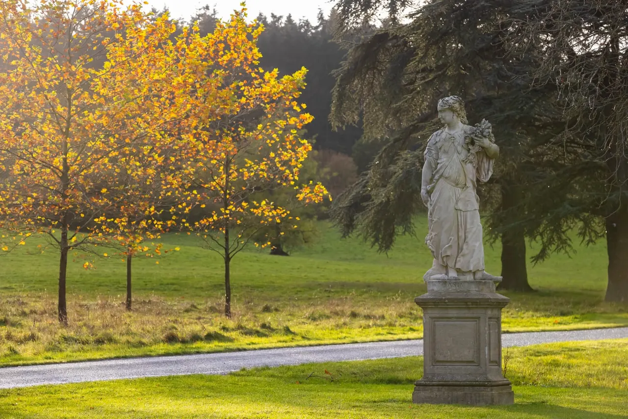 Stone statue of a classical female figure on a plinth in autumn parkland, with golden trees and evergreens nearby.