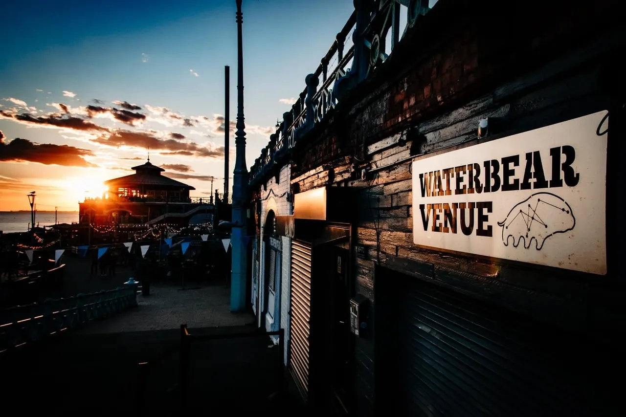 Waterfront pier at sunset with a sign for the venue, historic buildings, and golden sky reflecting on the water.