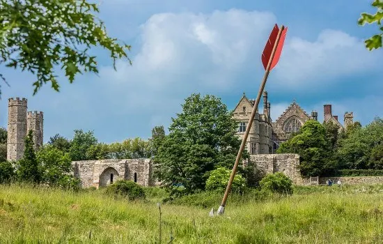 Giant arrow planted in grass field with medieval stone ruins and abbey buildings behind it.