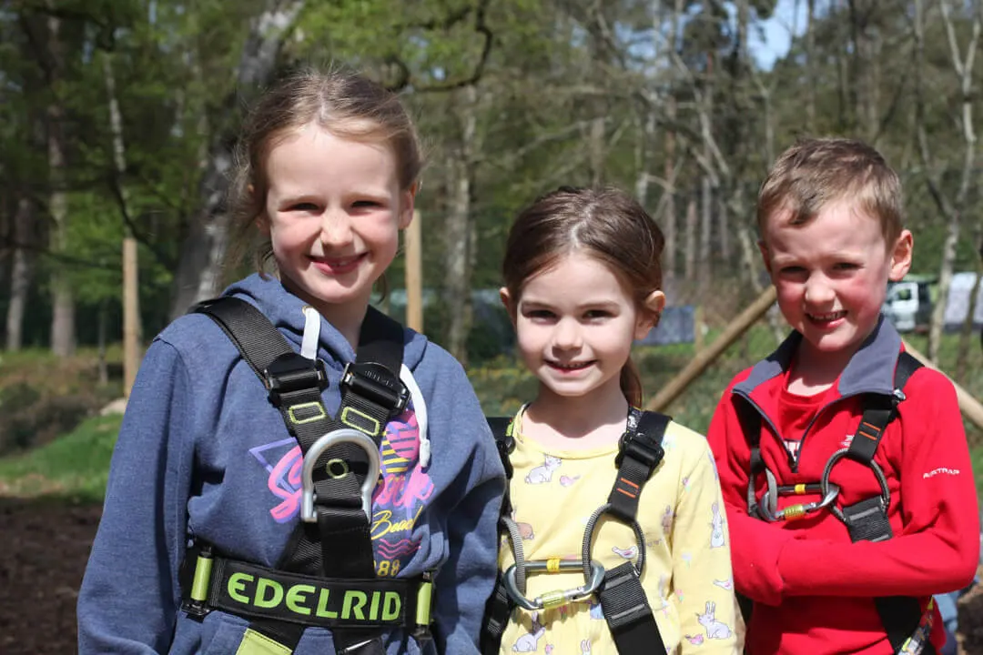 Three children wearing safety harnesses smile at the camera in a woodland setting.