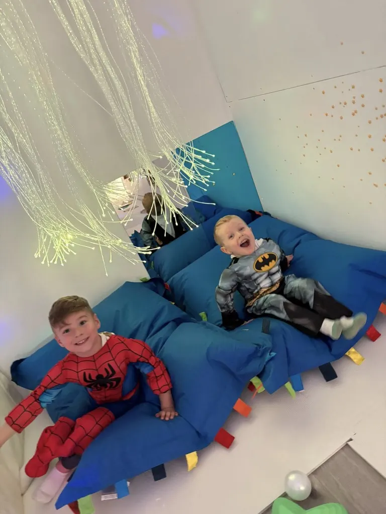 Two young children in superhero costumes lying on a blue padded play structure beneath hanging illuminated fibres.