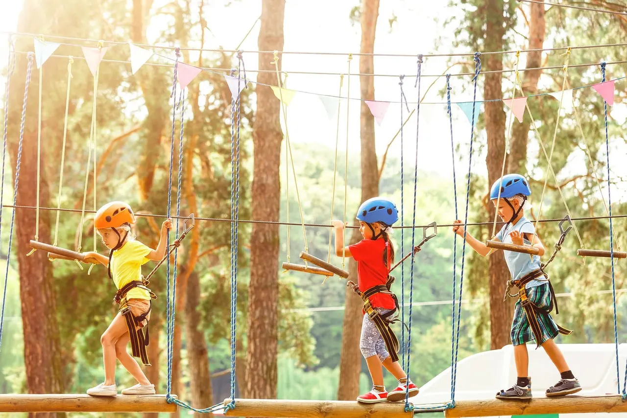 Three children wearing safety helmets and harnesses navigate a rope course through woodland, holding onto wooden logs.