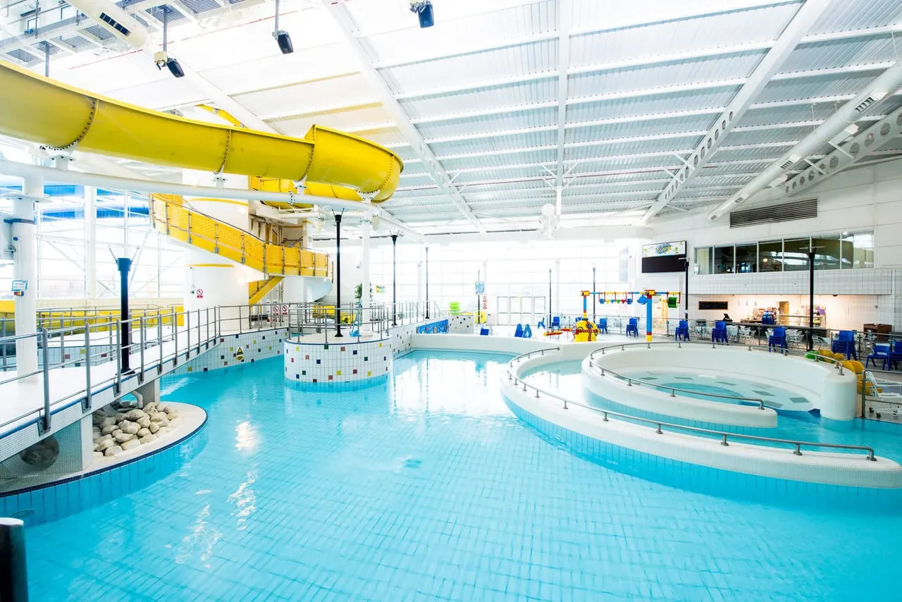 Indoor leisure centre with multiple swimming pools, a yellow water slide, diving platform, and a few visitors in the bright, modern facility.