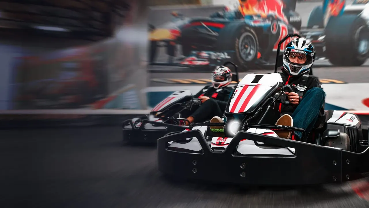 A person in a helmet driving a red and white go-kart on an indoor track with other karts racing in the background.