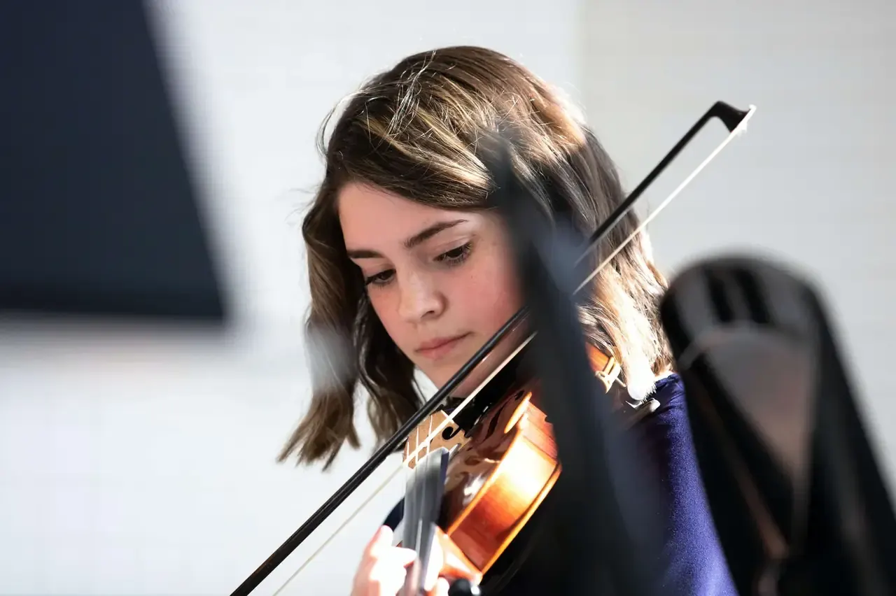 A young musician in a blue top plays a violin, looking down at the instrument during practice.