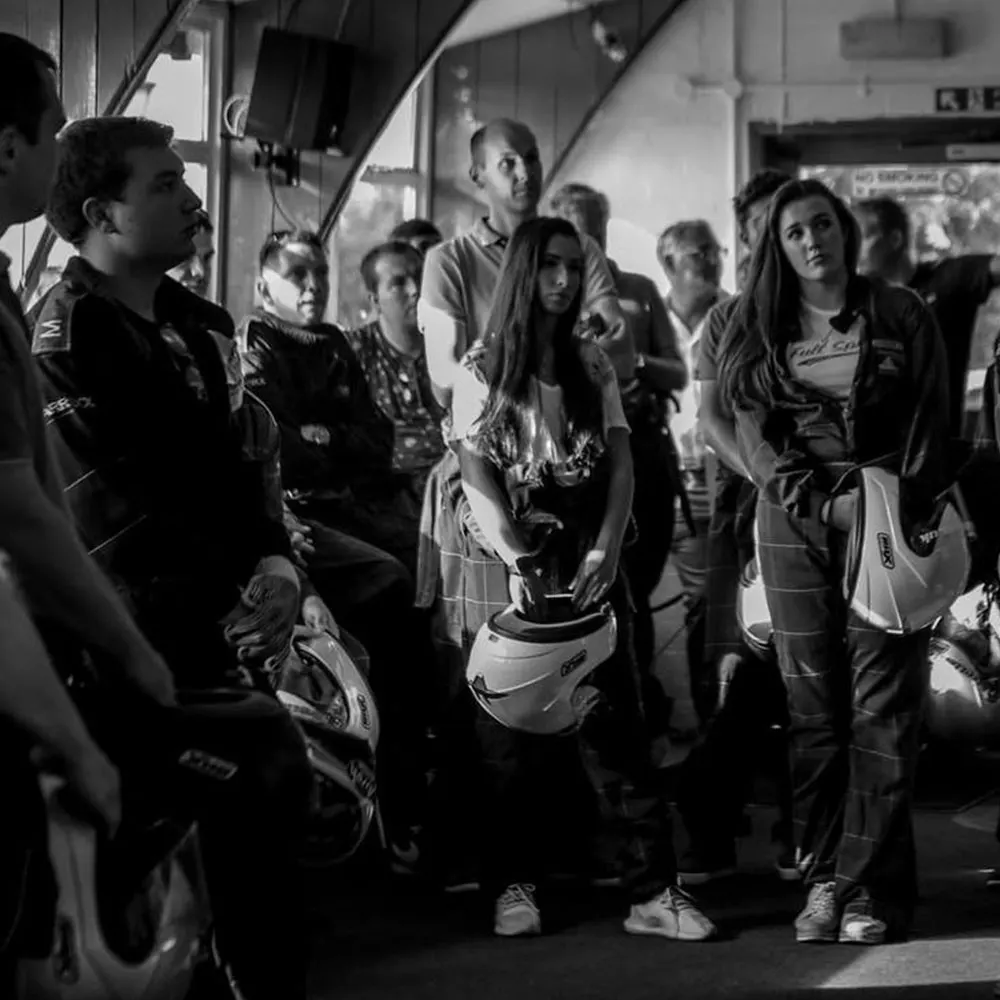 Group of people in racing suits and helmets gathered inside a pit garage or briefing area.