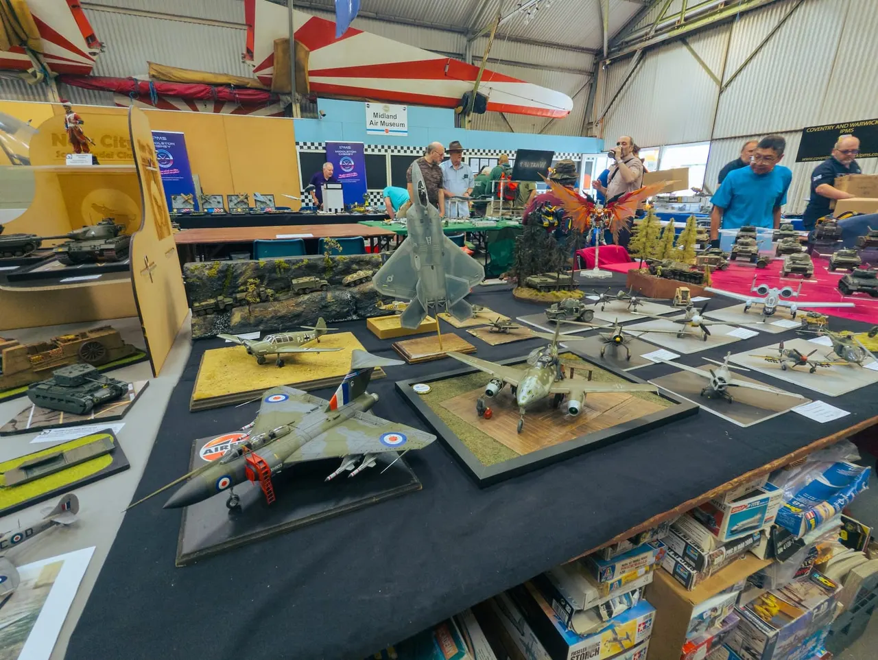 Scale model aircraft and military vehicles displayed on tables inside a hangar, with visitors browsing the exhibition.