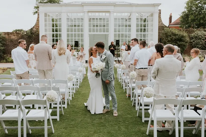 Wedding ceremony on lawn with bride and groom at altar, guests in white chairs, glasshouse and brick wall behind.