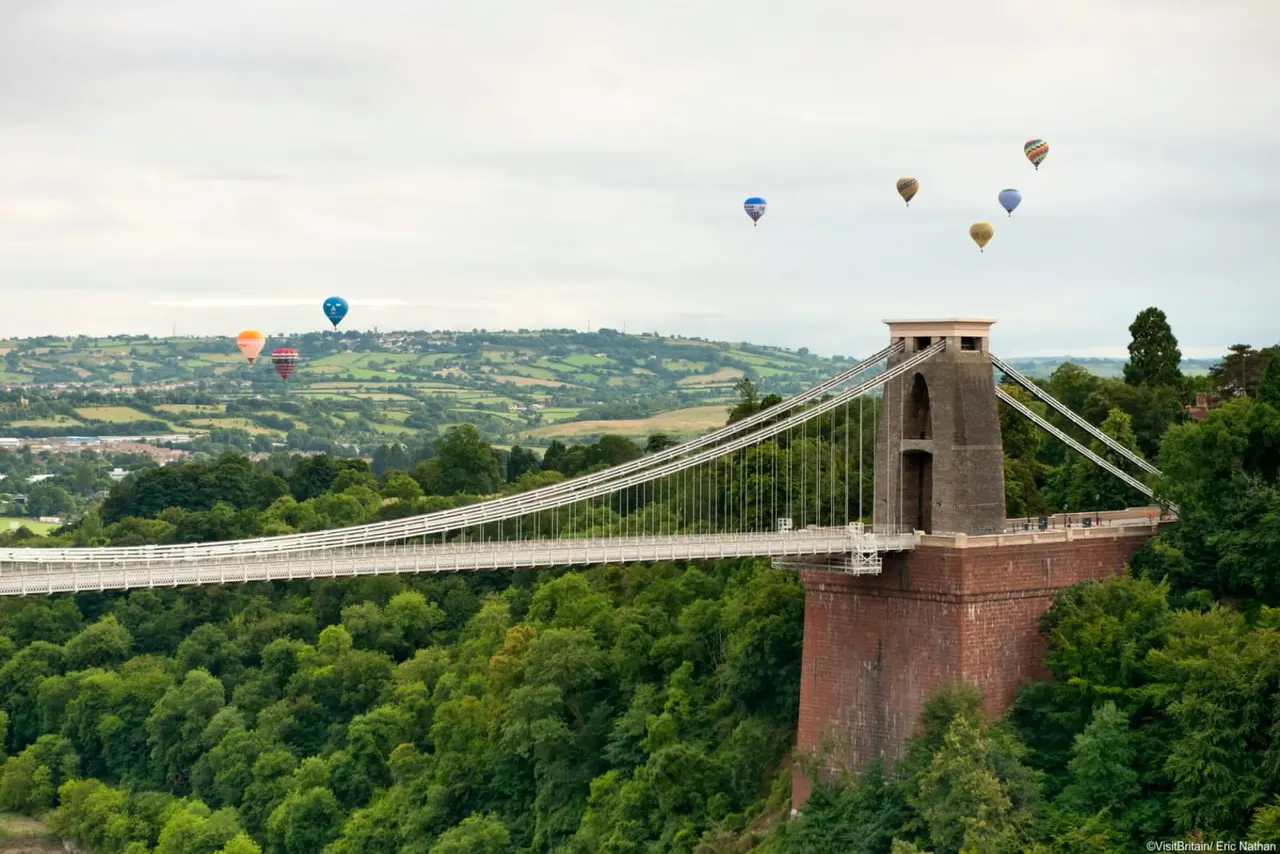Multiple hot air balloons float above the Clifton Suspension Bridge spanning a deep gorge with forested sides and rolling countryside beyond.