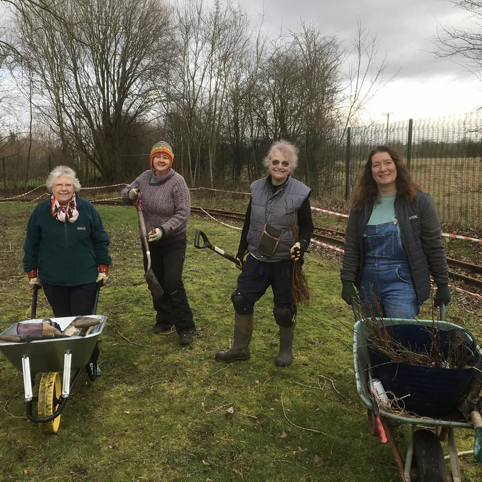 Four people posing with gardening tools in a vegetable plot with bare trees and fencing behind them.