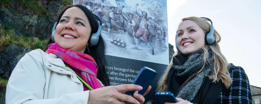 Two visitors wearing headphones hold a mobile device whilst looking at a historical information panel outdoors.